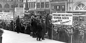 black and white photo of a demonstration.  One placard says, 'Compulsory vaccination German born - down with compulsion!', another says, 'Stop the slaughter of the innocents! Protest against compulsory vaccination.